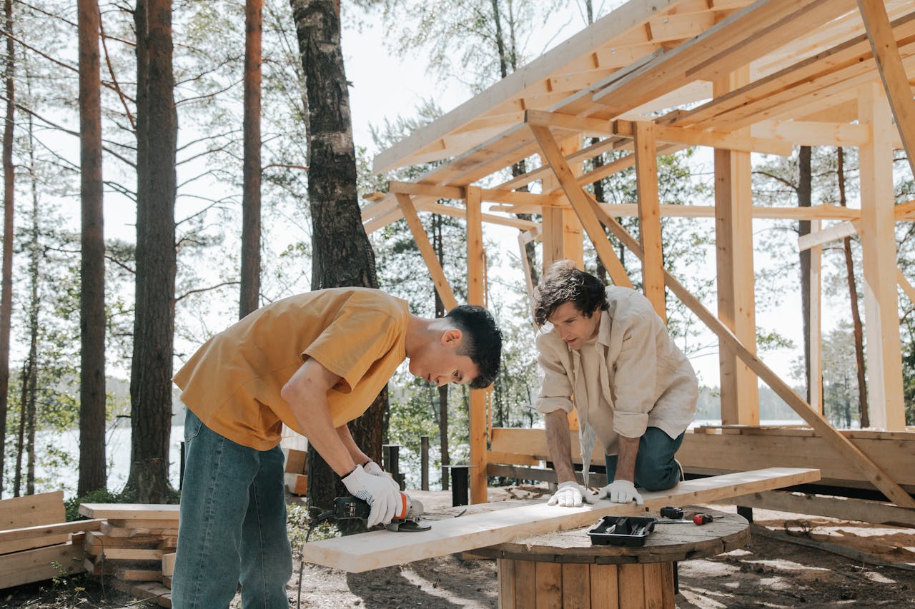Two carpenters working outdoors constructing a wooden frame in a forest setting.
